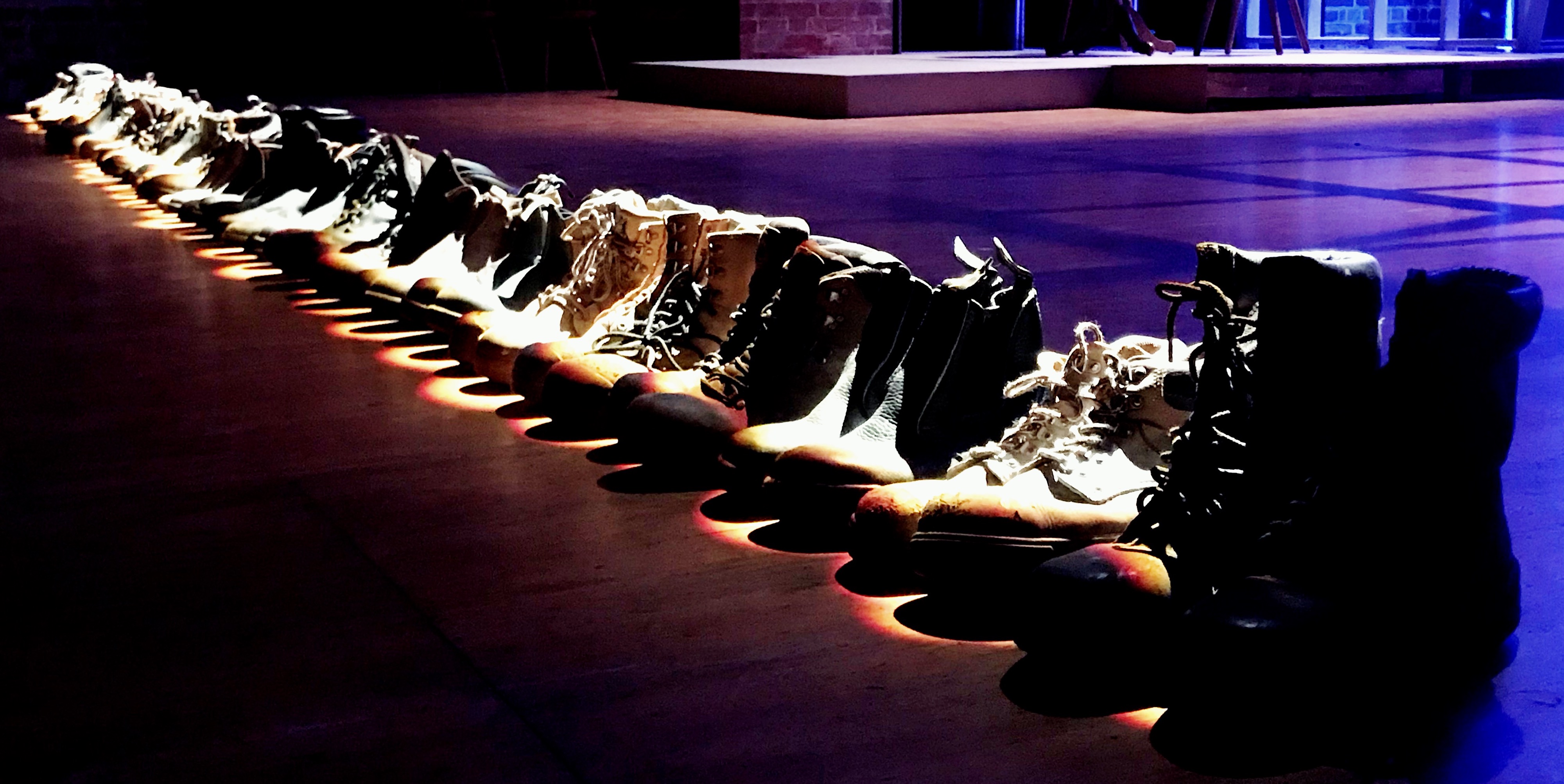 Photo by Elizabeth Humphrys: Final scene of the play 'The Bridge'. 35 pairs of boots for the 35 workers killed in the disaster, placed in a line across the front of the stage.