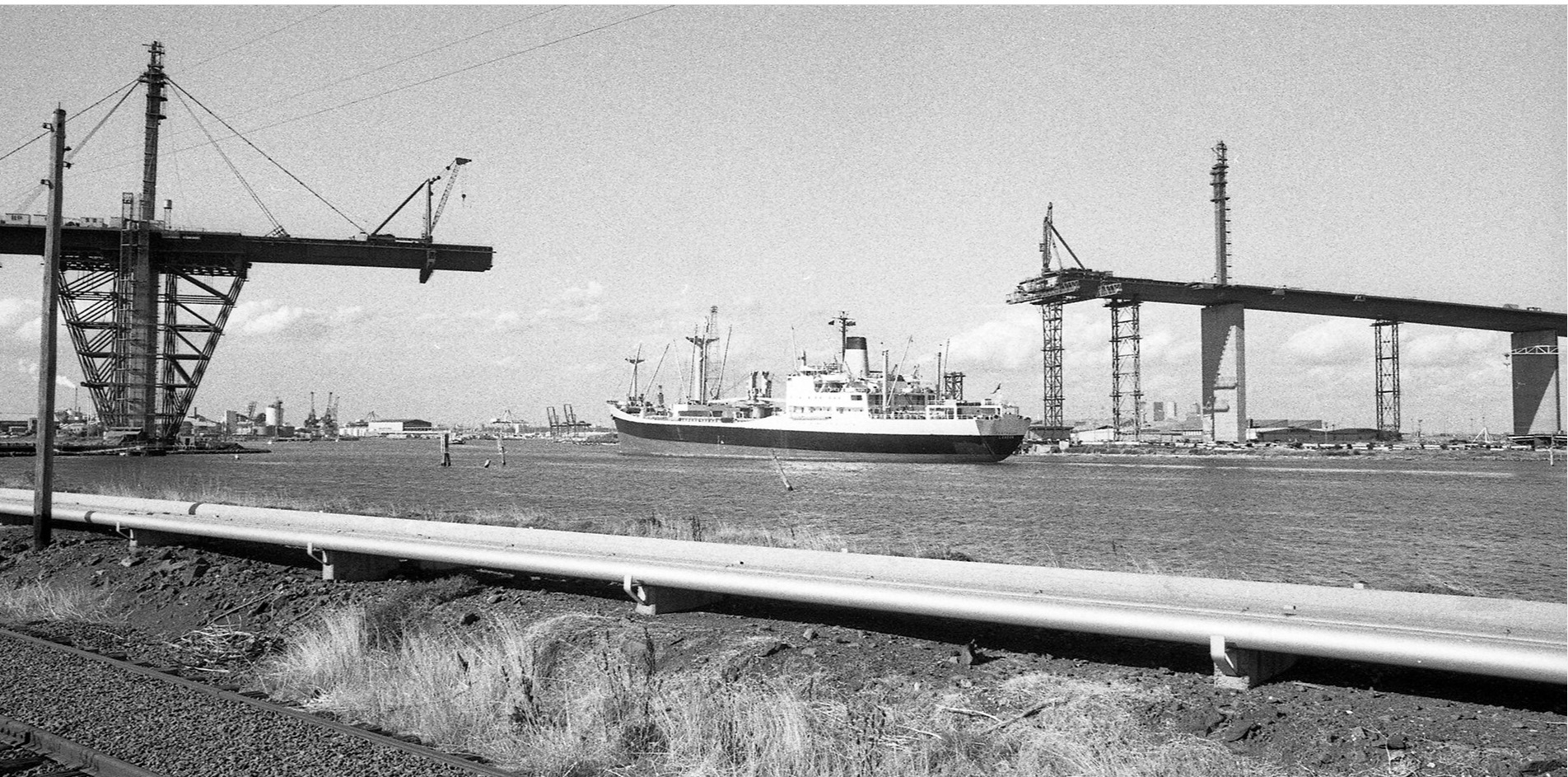 Partially finished West Gate, with spans from the west and east yet to meet in the midd.e Photo includes a large ship and construction cranes.
